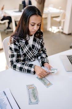 Female in plaid coat counting US dollar bills at office with laptop.