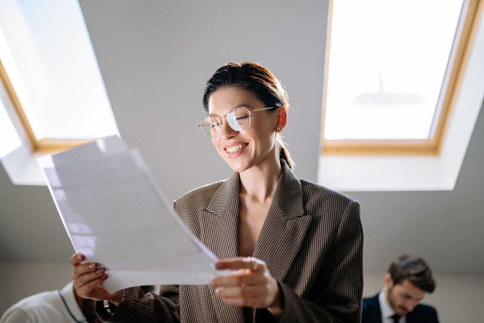 Smiling woman in glasses and a tweed blazer reading a printed document beneath bright skylights