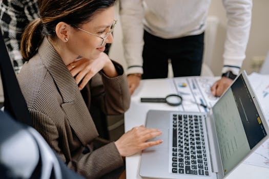 Businesswoman using laptop at office desk surrounded by colleagues, collaborating on project.