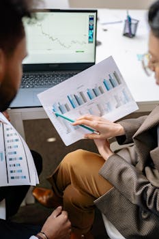 Two adults discuss financial data with charts and laptop in office setting.