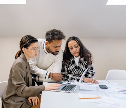 Three professionals collaborating at a desk with a laptop and documents, showcasing teamwork and diversity.