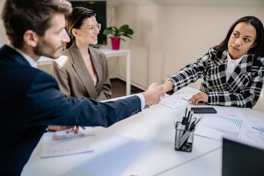 Three colleagues in a business meeting shaking hands over charts and documents.