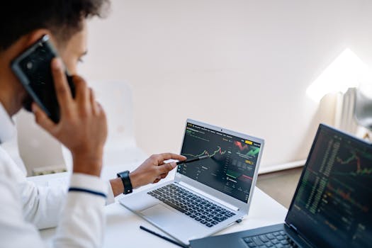 Business professional talking on phone and analyzing financial charts on laptops in office setting.