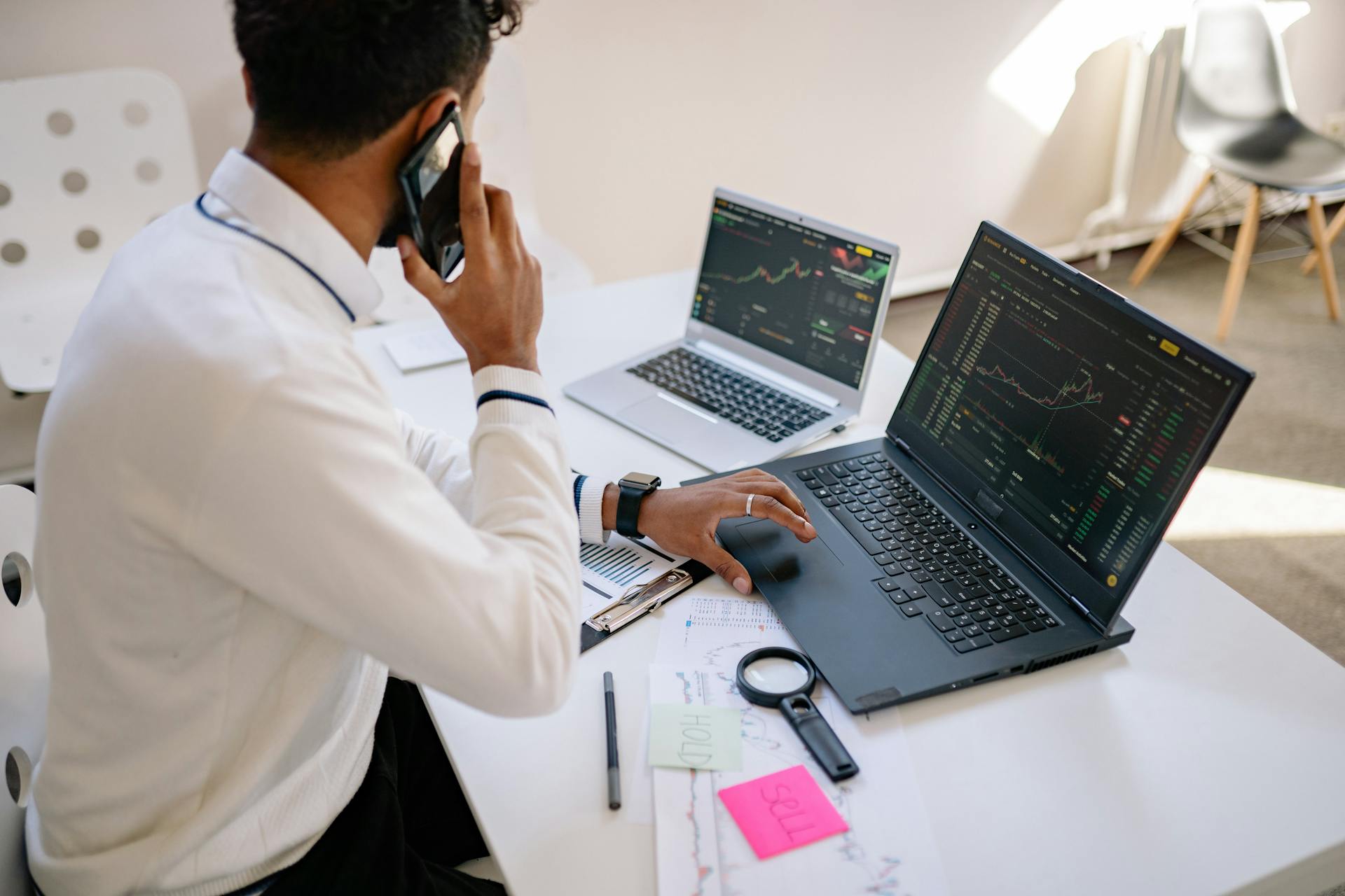 Man holding phone, working on multiple laptops with financial charts at desk.