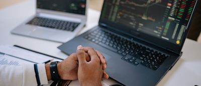 A person sitting at a desk with clasped hands, reviewing financial market charts on two laptop screens.