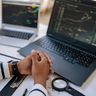 A person sitting at a desk with clasped hands, reviewing financial market charts on two laptop screens.