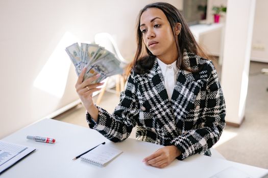 A young woman in a plaid coat counting US dollar bills at a desk in a modern office.