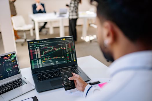 A businessman using smartphone and laptop to track stock market trends in a modern office setting.