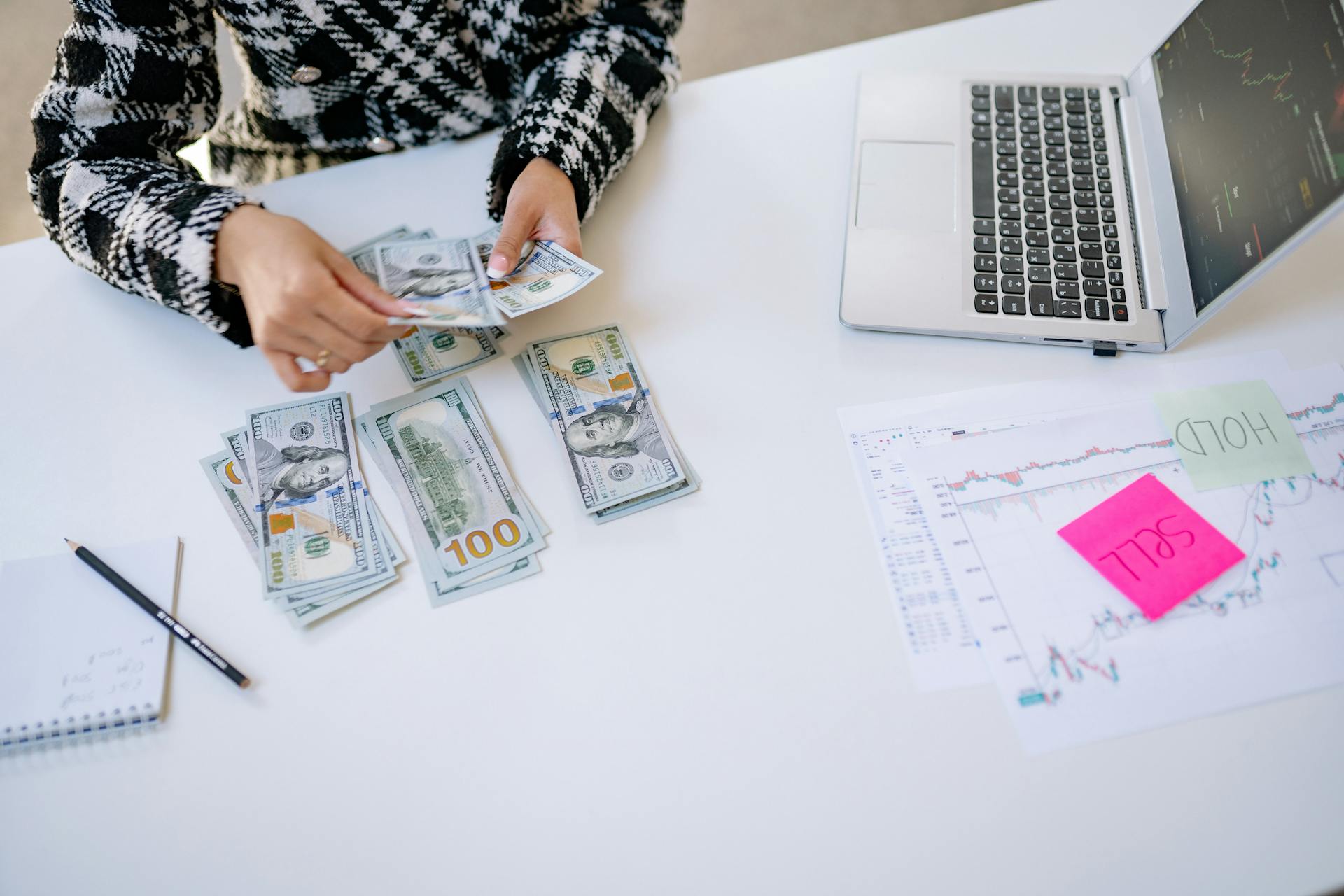 Person counting cash next to laptop and stock market charts on a white table.