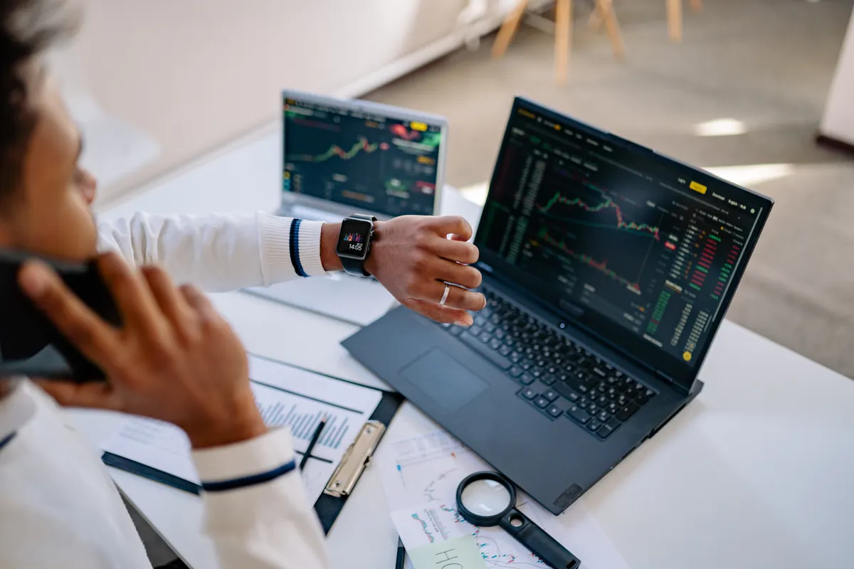 A trader checks a smartwatch while two laptops show futures charts, representing monitored semi-automated trading on a Blue Guardian account.