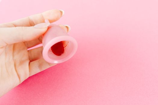 A hand holding a menstrual cup with blood against a pink background. Health and hygiene concept.