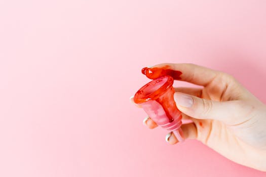 Close-up of a hand holding a menstrual cup with blood on a pink background.