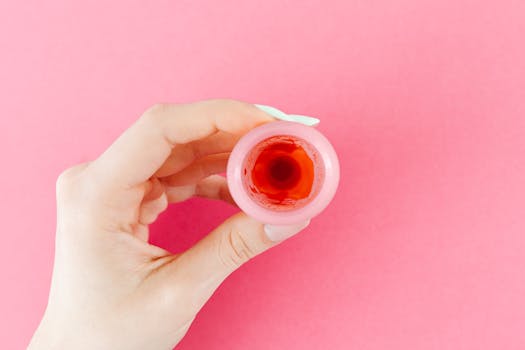 A detailed view of a menstrual cup with red liquid held by a hand against a pink backdrop, symbolizing menstruation.