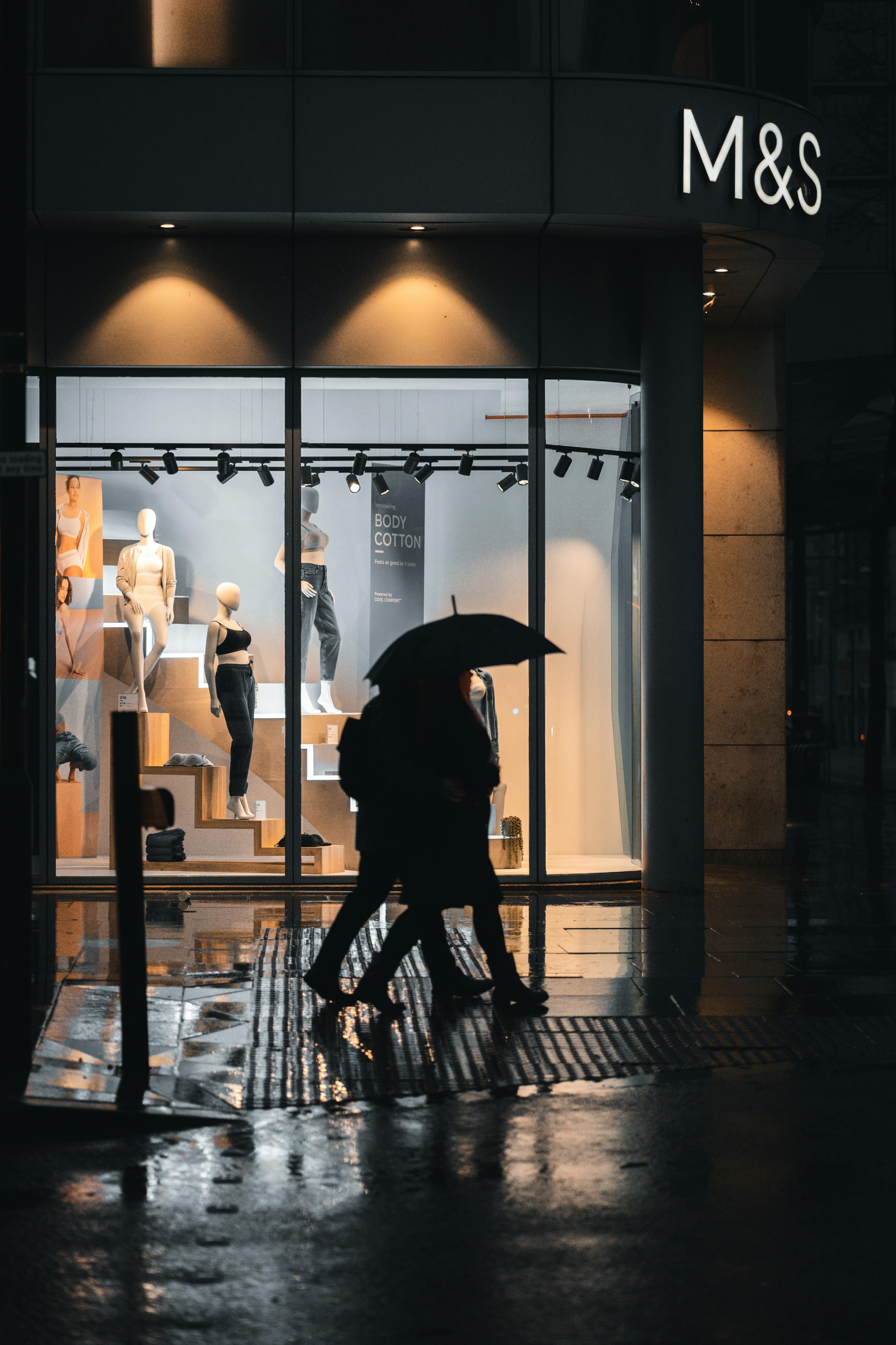 Silhouettes walking with umbrellas by a lit storefront on a rainy Manchester evening.