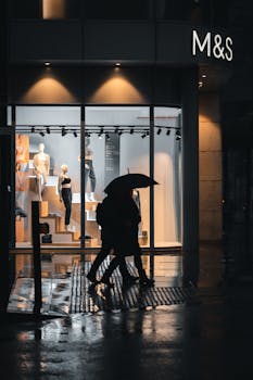 Silhouettes walking with umbrellas by a lit storefront on a rainy Manchester evening.