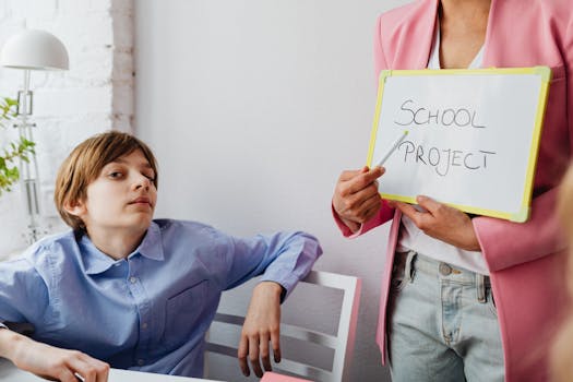 A teacher and student engage in a school project discussion indoors with a whiteboard.