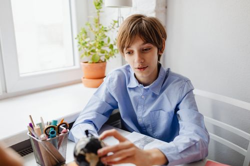 Free Teenager in blue shirt studying at a desk with a globe and stationery indoors near a window. Stock Photo