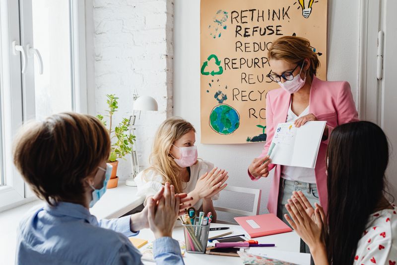 Professor e alunos com máscaras discutindo um projeto escolar criativo e ecológico em um ambiente interno.