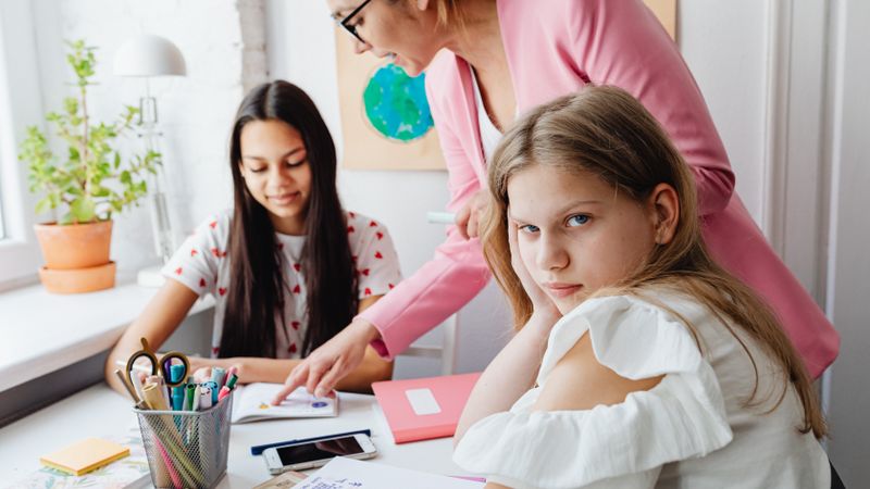 A teacher works alongside two students on schoolwork at a desk in a bright classroom