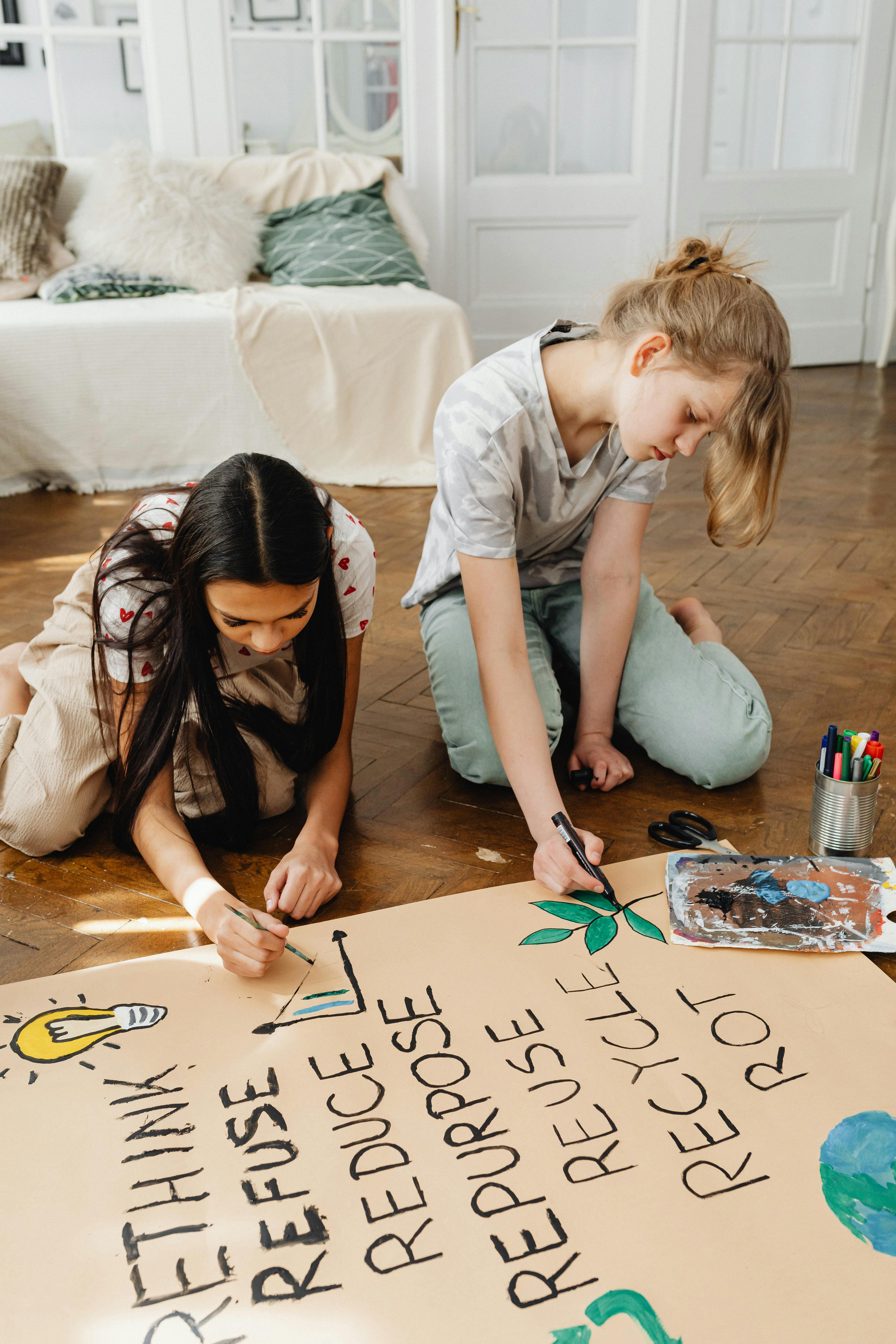 Two girls engaged in creating a recycling-themed poster on the floor indoors.