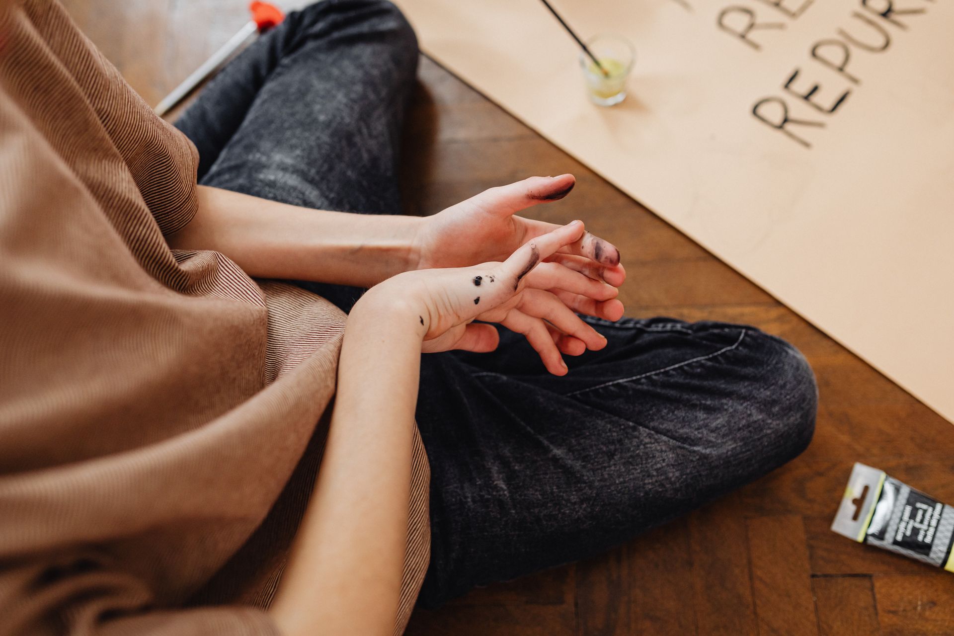 Close-up view of a person sitting on floor with paint-stained hands, creating art.