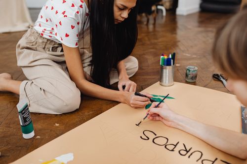 Free Two young adults engaged in a creative art project indoors, painting a poster with brushes and markers. Stock Photo