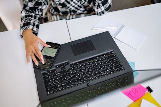 A person working with a smartphone and laptop on a white desk in an office setting.