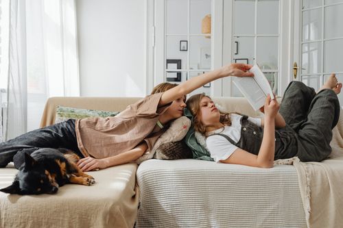 Free Two siblings lounging on a couch, reading together, with a dog resting beside them. Stock Photo