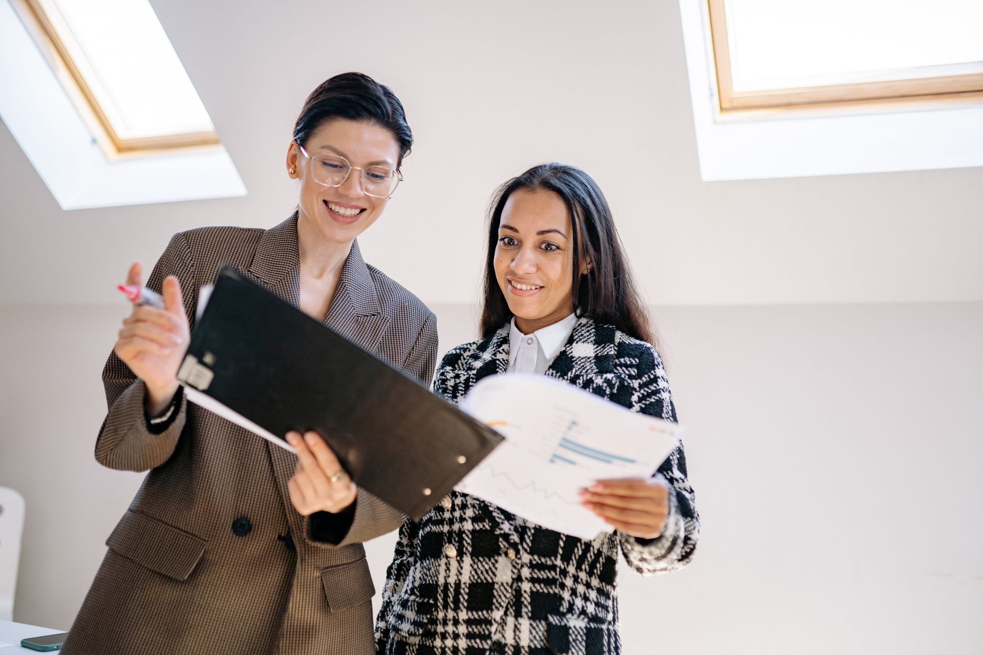 Two professional women in business attire smiling and reviewing documents indoors.