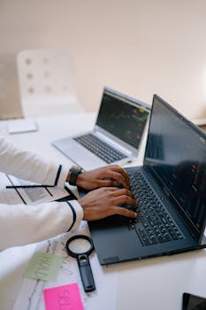 A person working on two laptops with financial charts, emphasizing remote productivity.