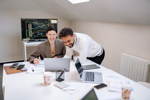 Two colleagues engaging in teamwork with laptops and documents, smiling in a modern office.