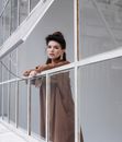 Attractive female in trendy outfit with black hair looking at camera while leaning on frame of window in modern building