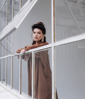 Attractive female in trendy outfit with black hair looking at camera while leaning on frame of window in modern building