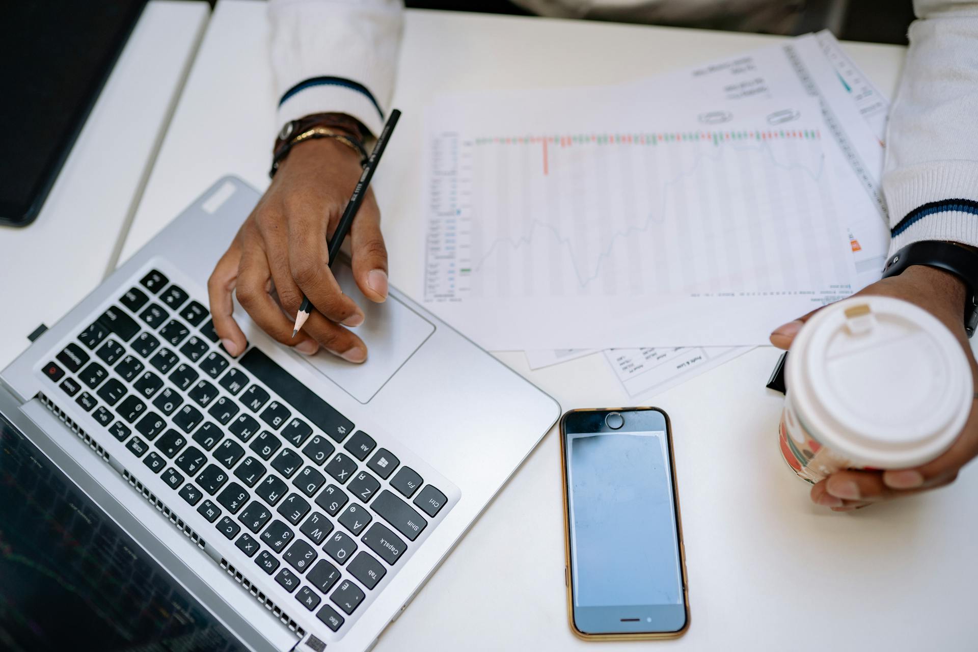 A businessman holding coffee and using a laptop at a desk with charts and a smartphone.