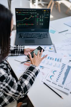 Businesswoman evaluating stock market trends using graphs and smartphone in office.