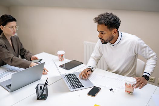 Two colleagues working together in an office with laptops, indicating teamwork and productivity.
