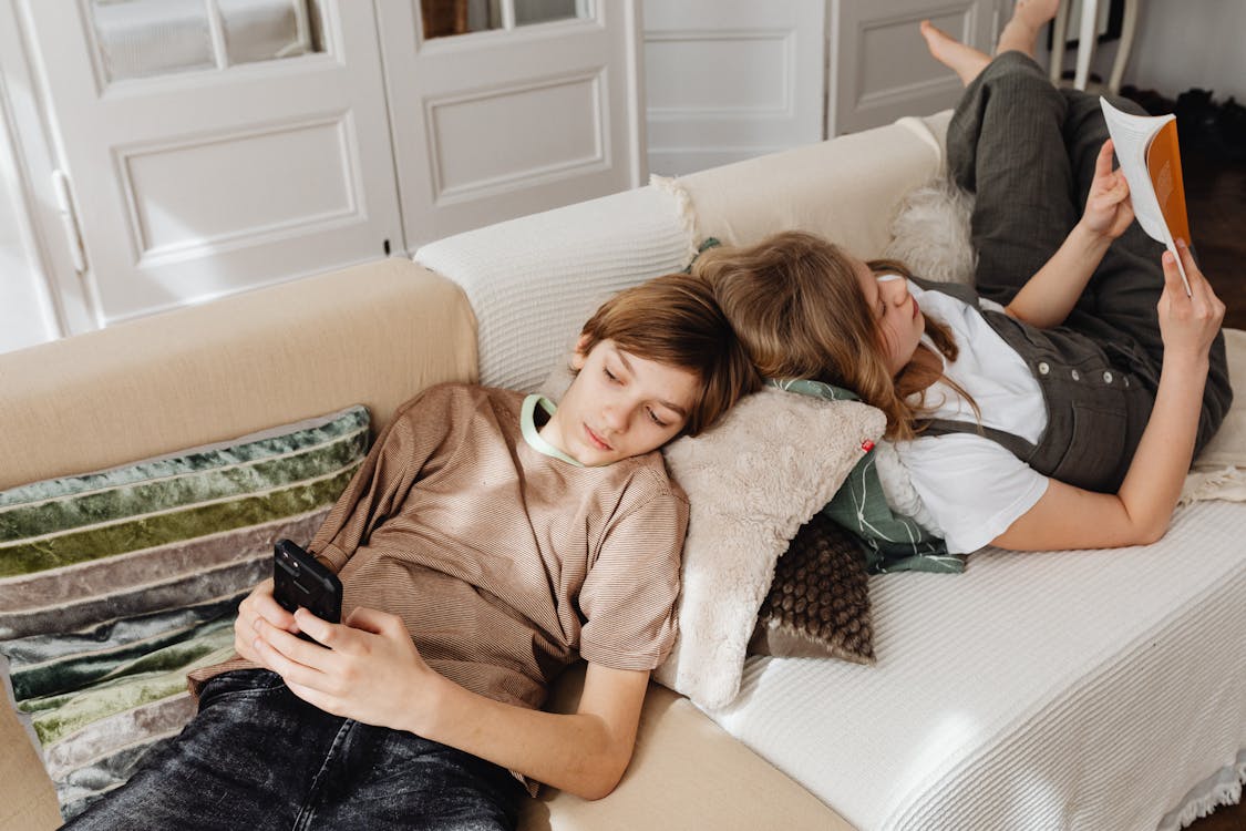 Free Teenagers lounging on the couch at home, browsing a phone and reading a book. Stock Photo