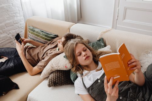 Free Siblings enjoying leisure time reading and using a phone on a cozy living room couch. Stock Photo