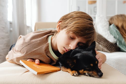 A young boy rests his head on a black dog while holding a book, depicting calmness and companionship.