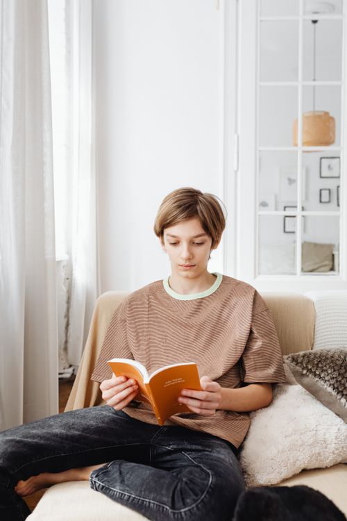 Free Teenager relaxing while reading a book in a cozy living room setting. Stock Photo