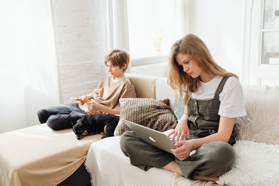 Two teenagers on a couch using a laptop and smartphone indoors with a pet dog present.