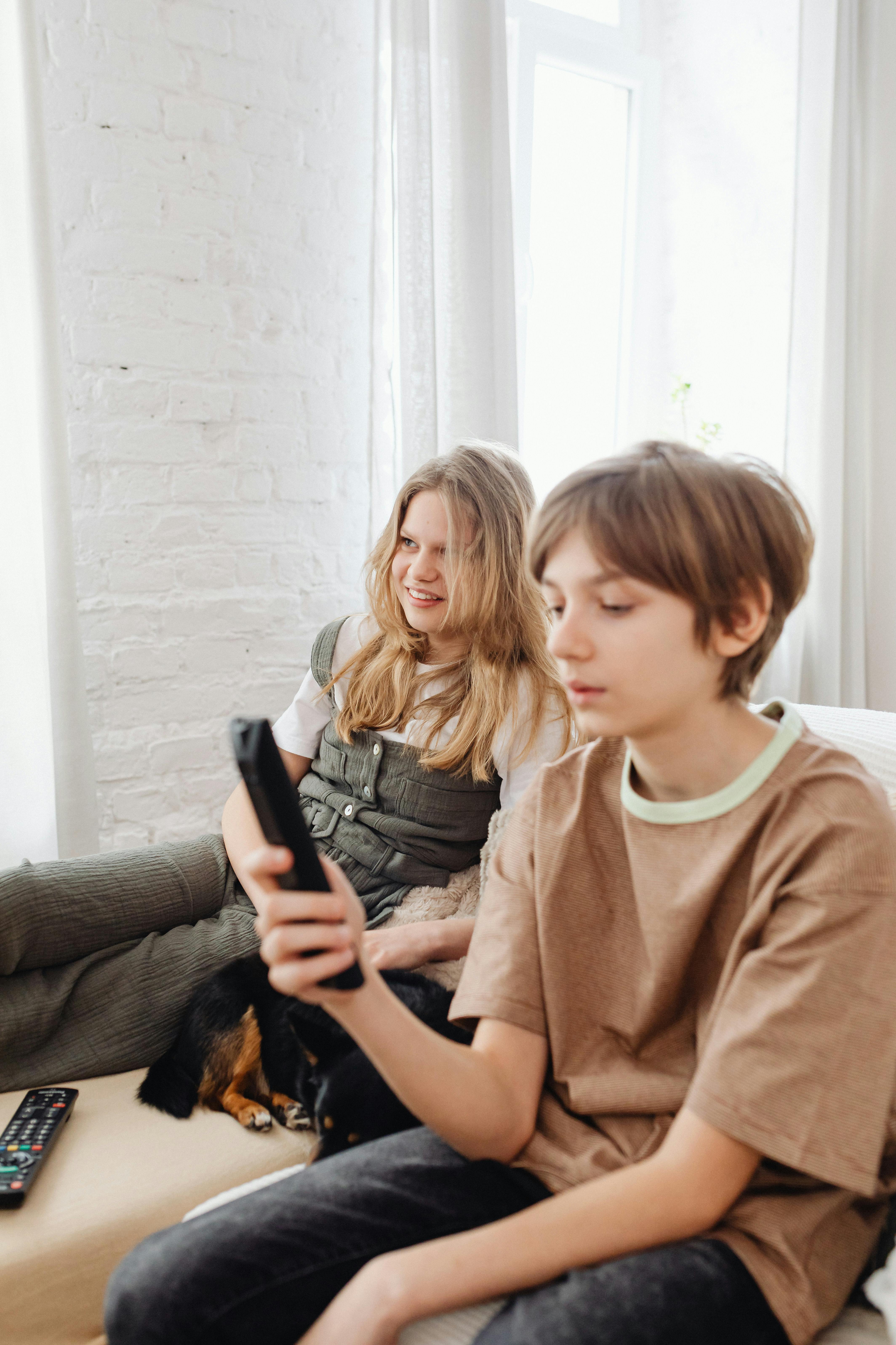 Two siblings sitting indoors with a dog, holding a TV remote and enjoying leisure time.