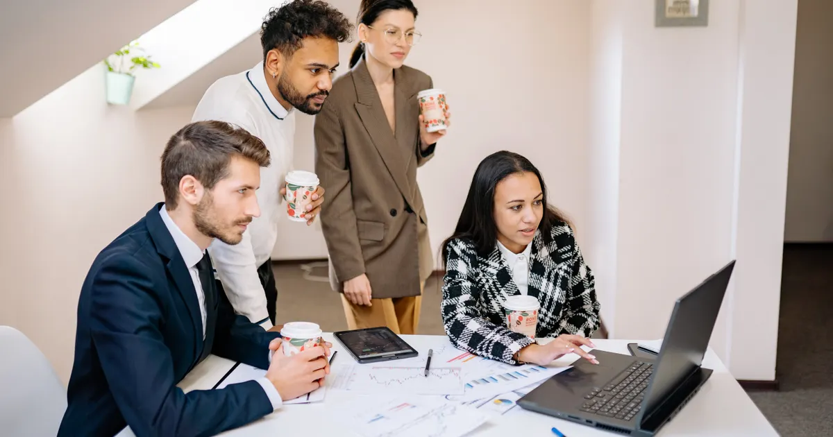 Four colleagues working together on a laptop around a table with coffee cups