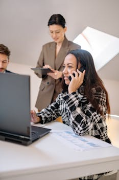 Businesswomen collaborating in an office, discussing strategies on phone and tablet.