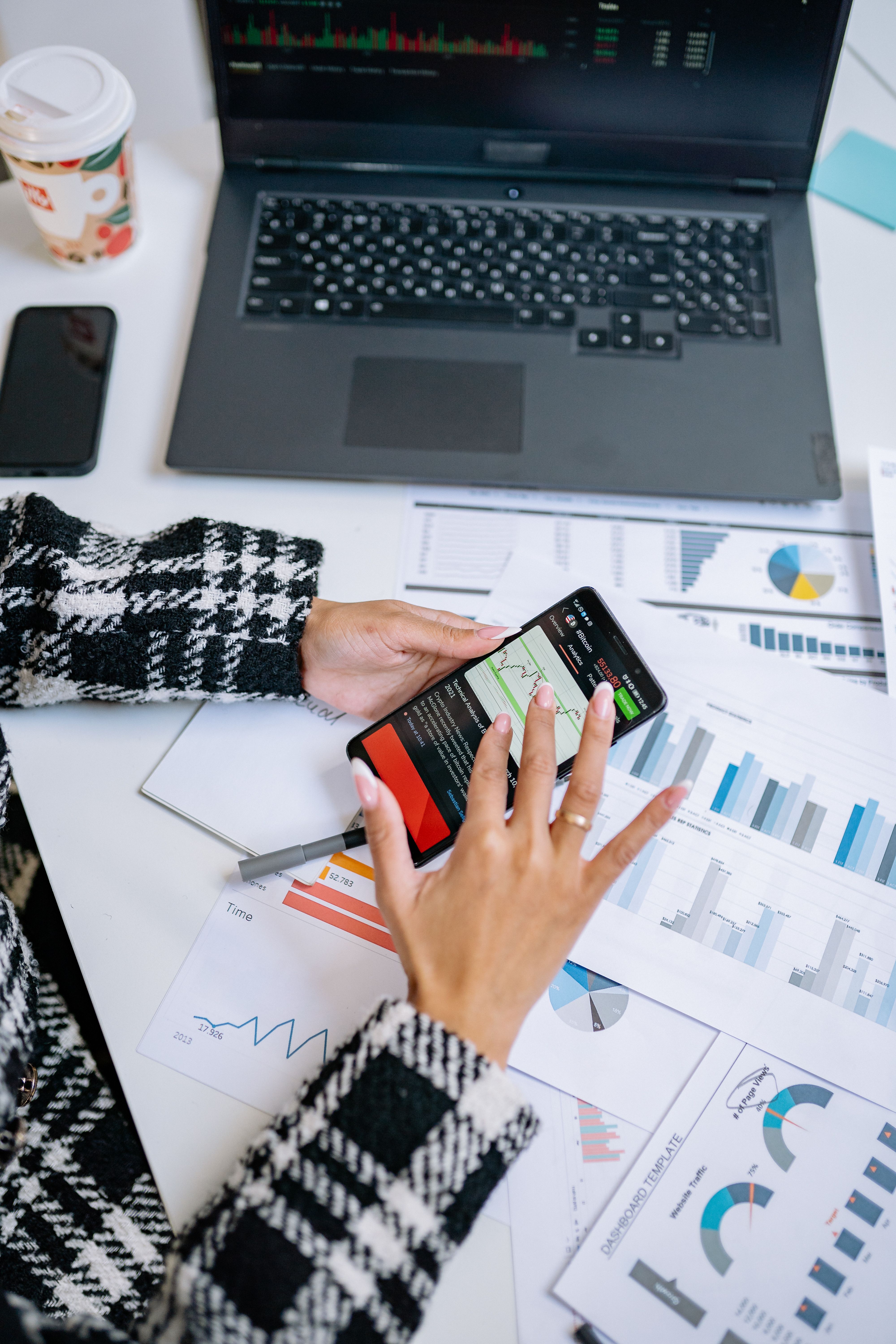 Pessoa analisando graficos e dados no smartphone ao lado de laptop em mesa de trabalho