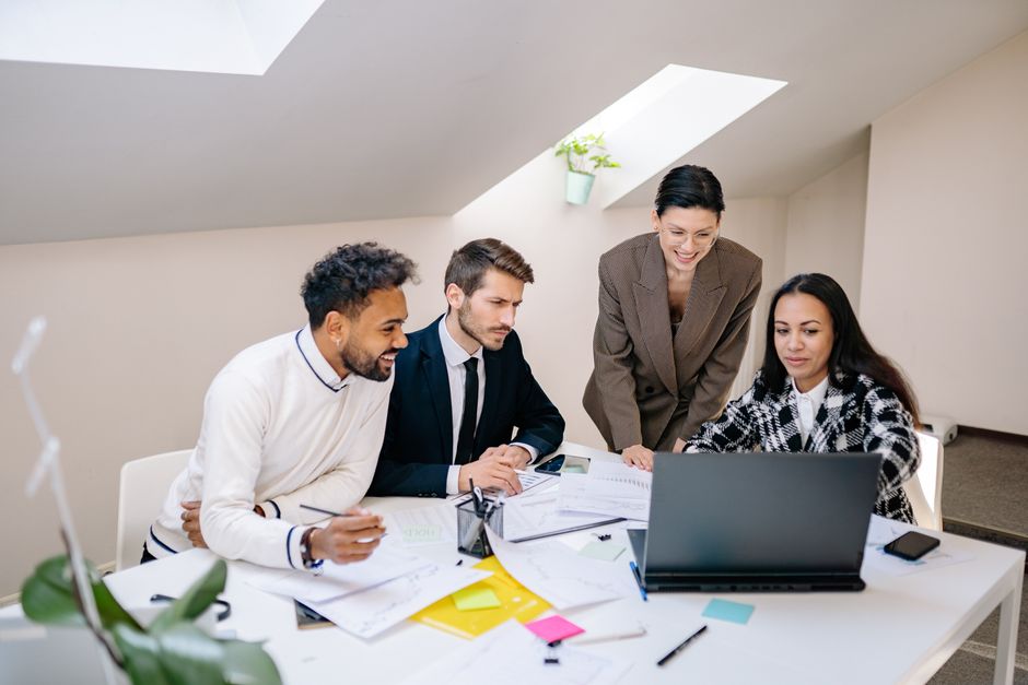 Four colleagues engaged in a collaborative meeting around a laptop in a bright office space.