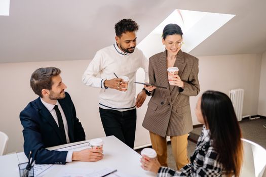 A diverse team of young professionals discussing a project over coffee in a modern office setting.