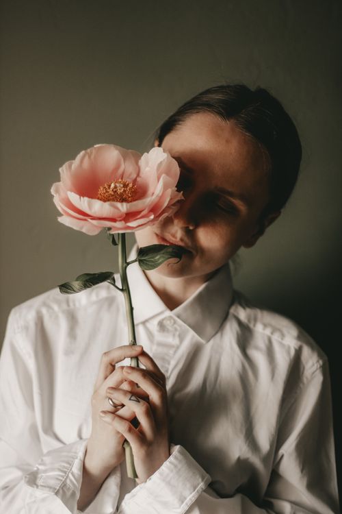 Free Calm woman in white blouse standing with closed eyes and blooming pink peony flower in hands against dark background in room Stock Photo