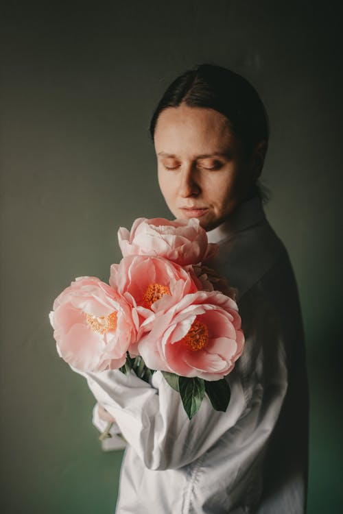Free Tranquil female in casual clothes standing with closed eyes and smelling bouquet of blooming pink peonies against dark wall in room in daytime Stock Photo