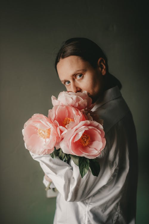 Free Side view of young female with dark hair in white blouse standing with bouquet of blooming peonies in room against dark background in daytime Stock Photo
